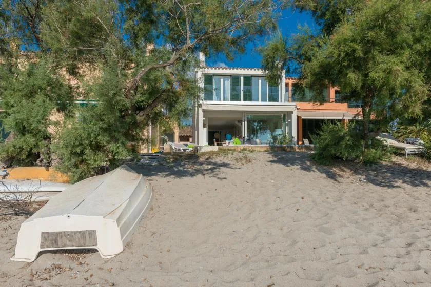 A modern beachfront house stands behind a sandy beach and leafy trees.