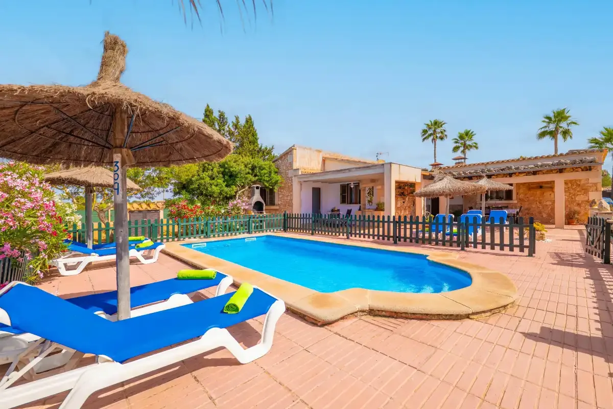 A swimming pool sits beside a tiled patio, shaded loungers, and a low building.