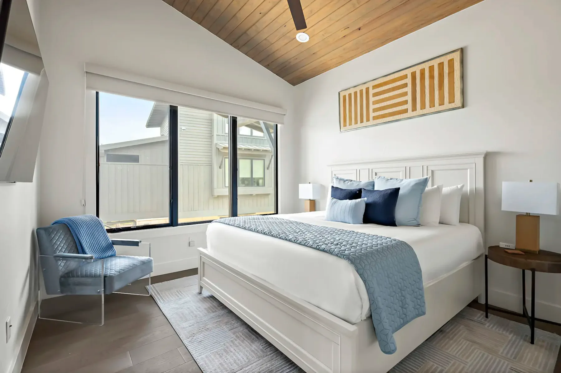Modern bedroom with a white bed, blue accents, large window, wood ceiling, and wall art. A small side table and chair complete the decor.