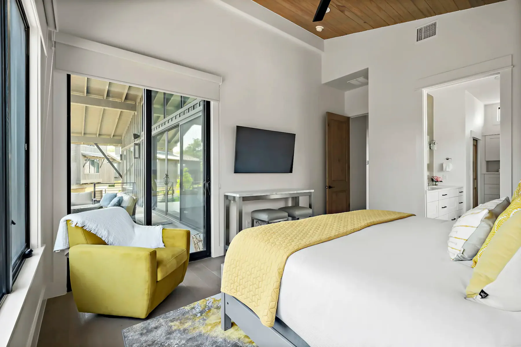 Bright bedroom with a large window, yellow armchair, wall-mounted TV, and an inviting bed with yellow accents next to a small desk.