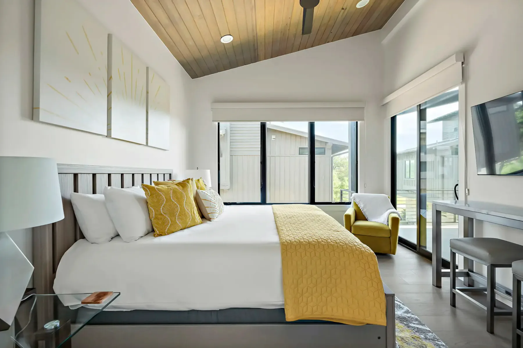 Modern bedroom with a large bed, yellow accents, abstract wall art, a wall-mounted TV, and large windows letting in natural light.