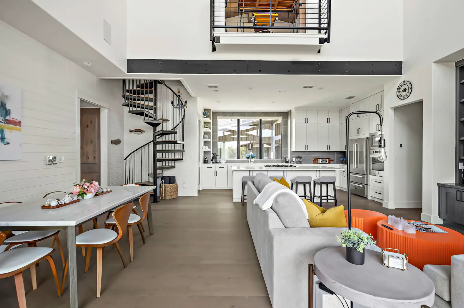 Modern living room with open kitchen, spiral staircase, gray sofa, orange chairs, and a dining table set. Bright and spacious interior.
