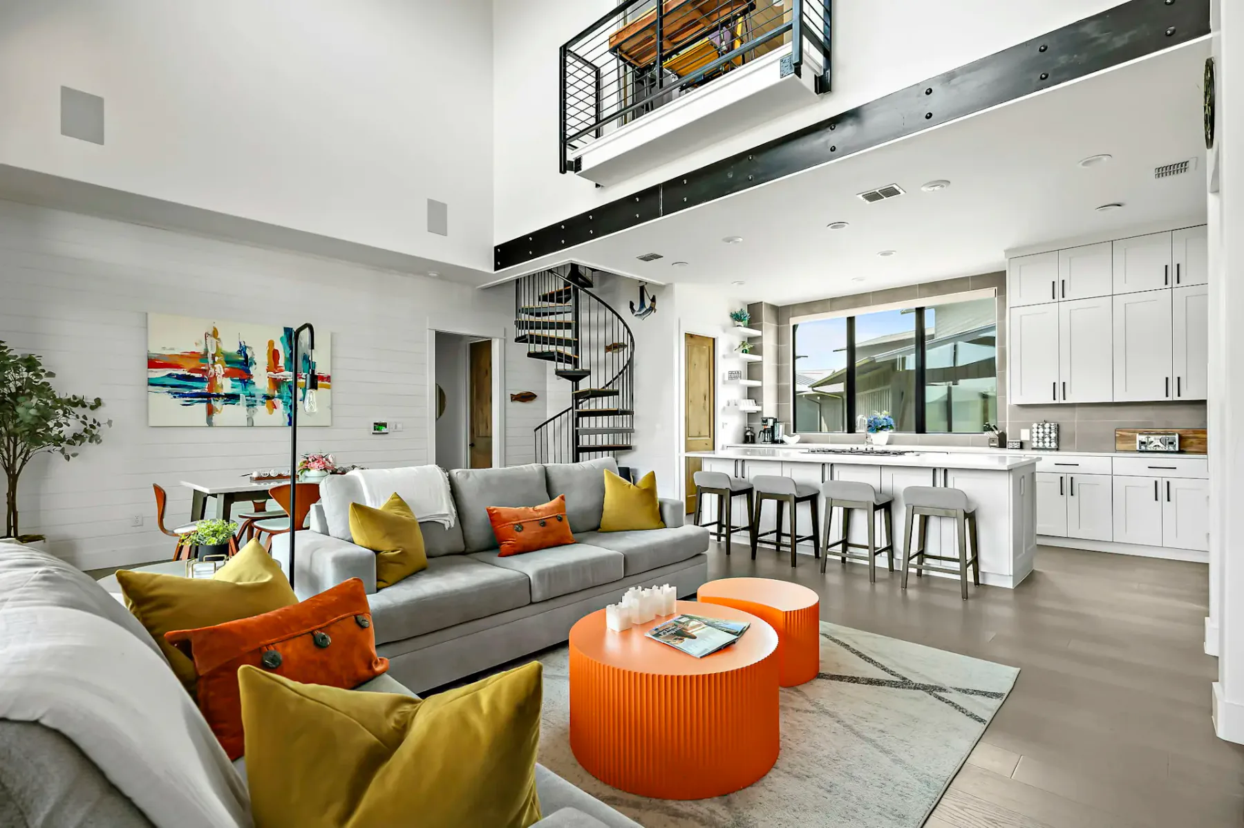 Modern open-concept living room with spiral staircase, gray sofas, orange accents, large window, and kitchen island with white cabinets.