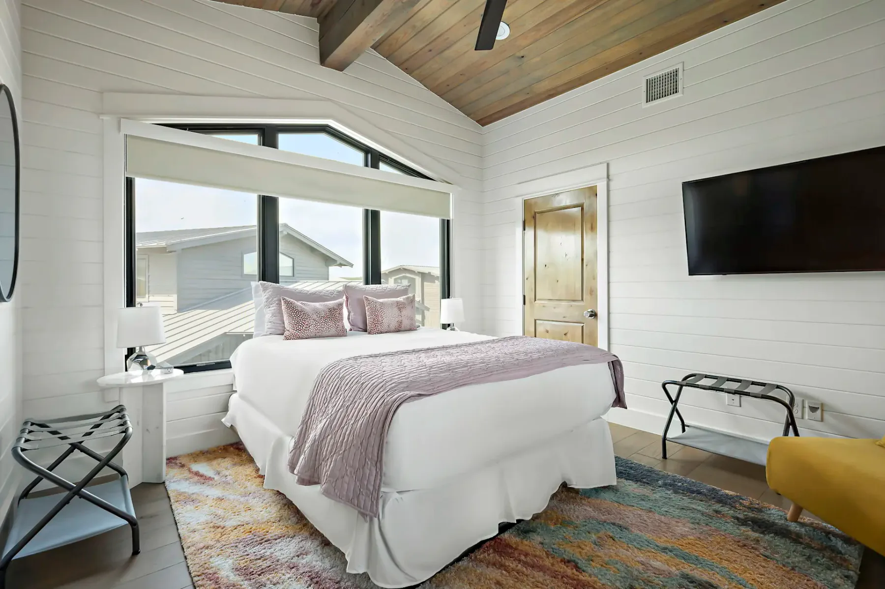 Bright bedroom with a large bed, pink pillows, wood ceiling, TV, and a colorful rug. Large window offers a view of rooftops.