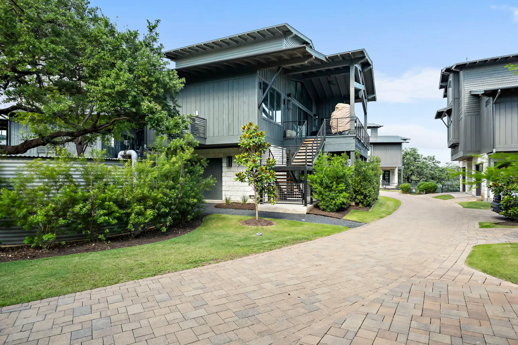 Modern house with grey facade, large windows, and a metal staircase, surrounded by greenery, on a paved path under a clear blue sky.