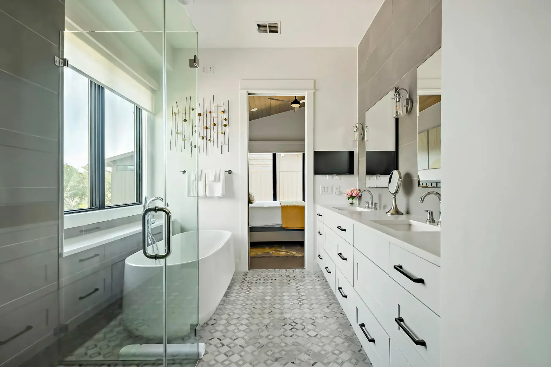 Modern bathroom with a glass-enclosed shower, freestanding tub by large windows, dual sinks on a white vanity, and geometric tiled floor.