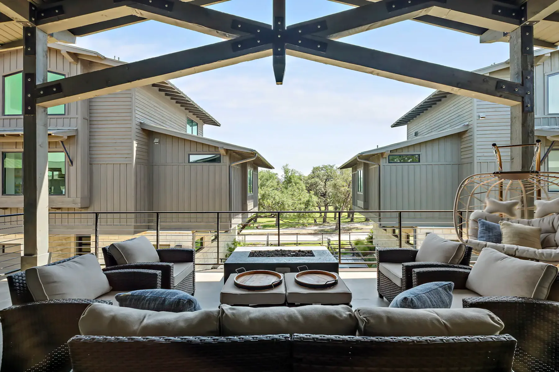 Outdoor patio with cushioned seating, a coffee table with plates, and hanging chairs, surrounded by modern buildings and greenery.
