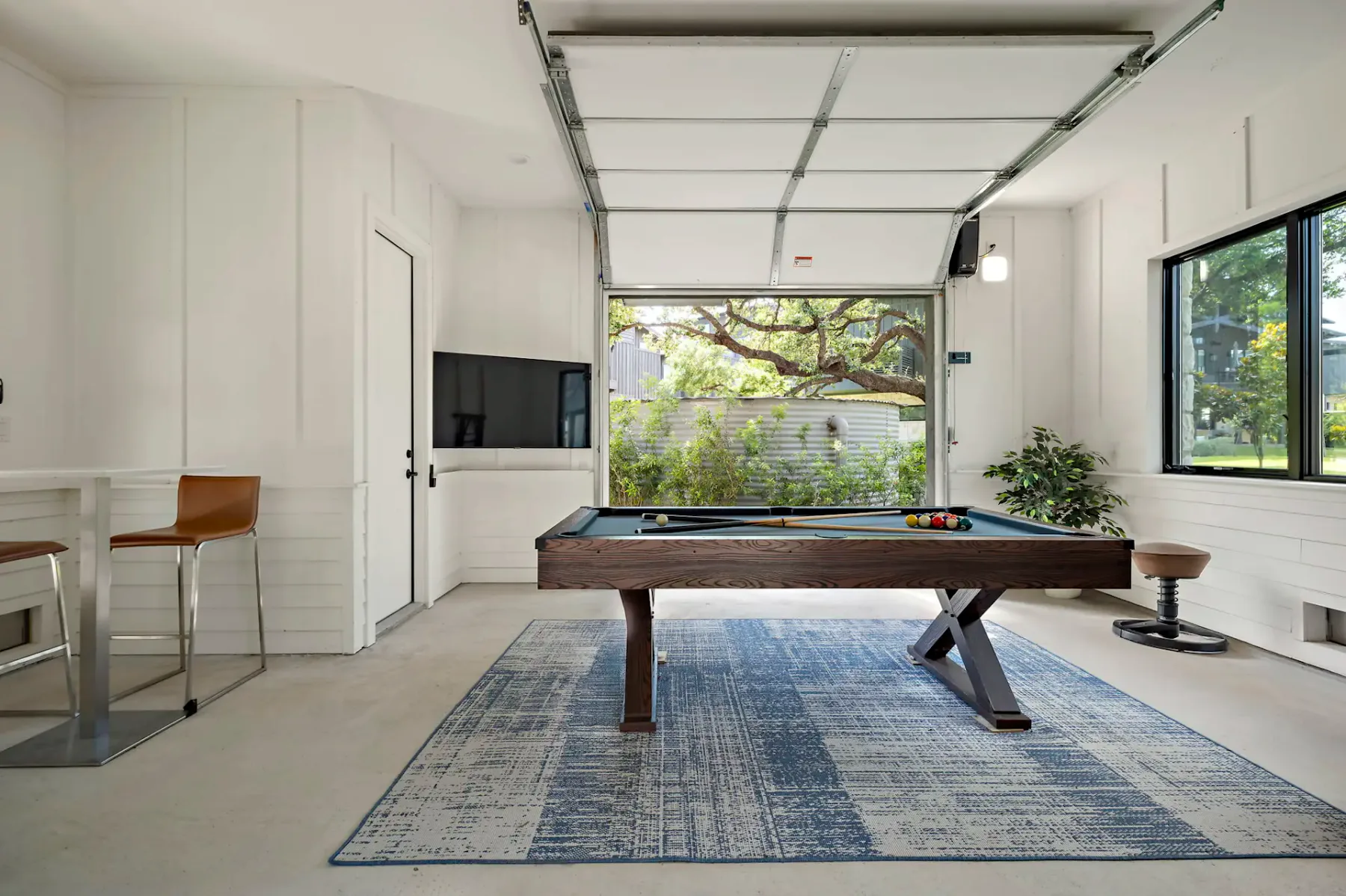 A pool table stands on a blue rug in a bright room with an open garage door.