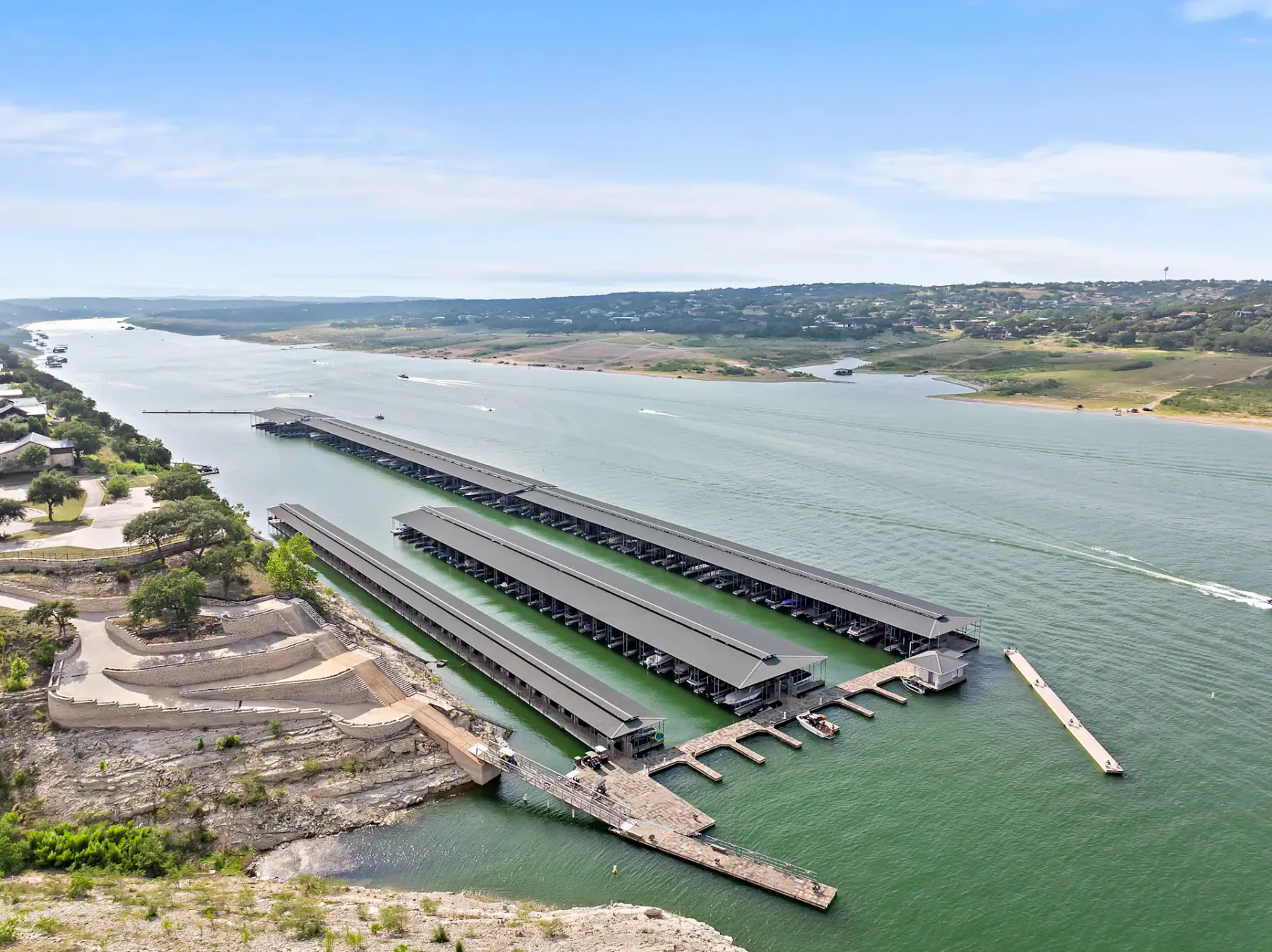 Aerial view of a marina with multiple docks and boat slips extending into a wide river, surrounded by rocky and grassy landscapes under a clear sky.