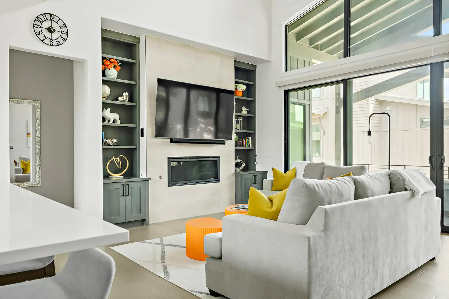 Modern living room with a large TV above a fireplace, gray sofa with yellow pillows, orange ottoman, and floor-to-ceiling windows.