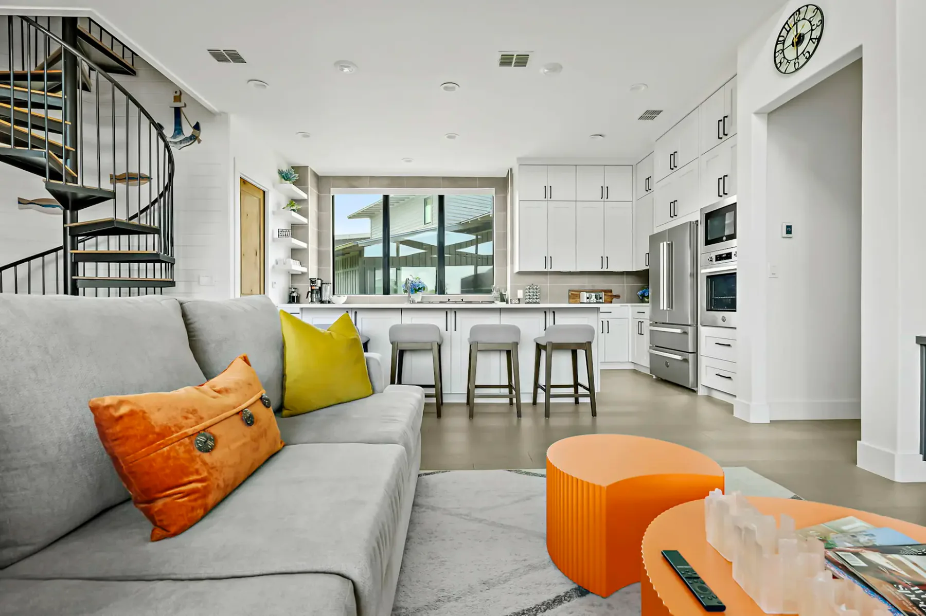 Modern living room with gray couch, orange tables, and kitchen. Spiral staircase on the left, clock on the wall, and large window in the background.