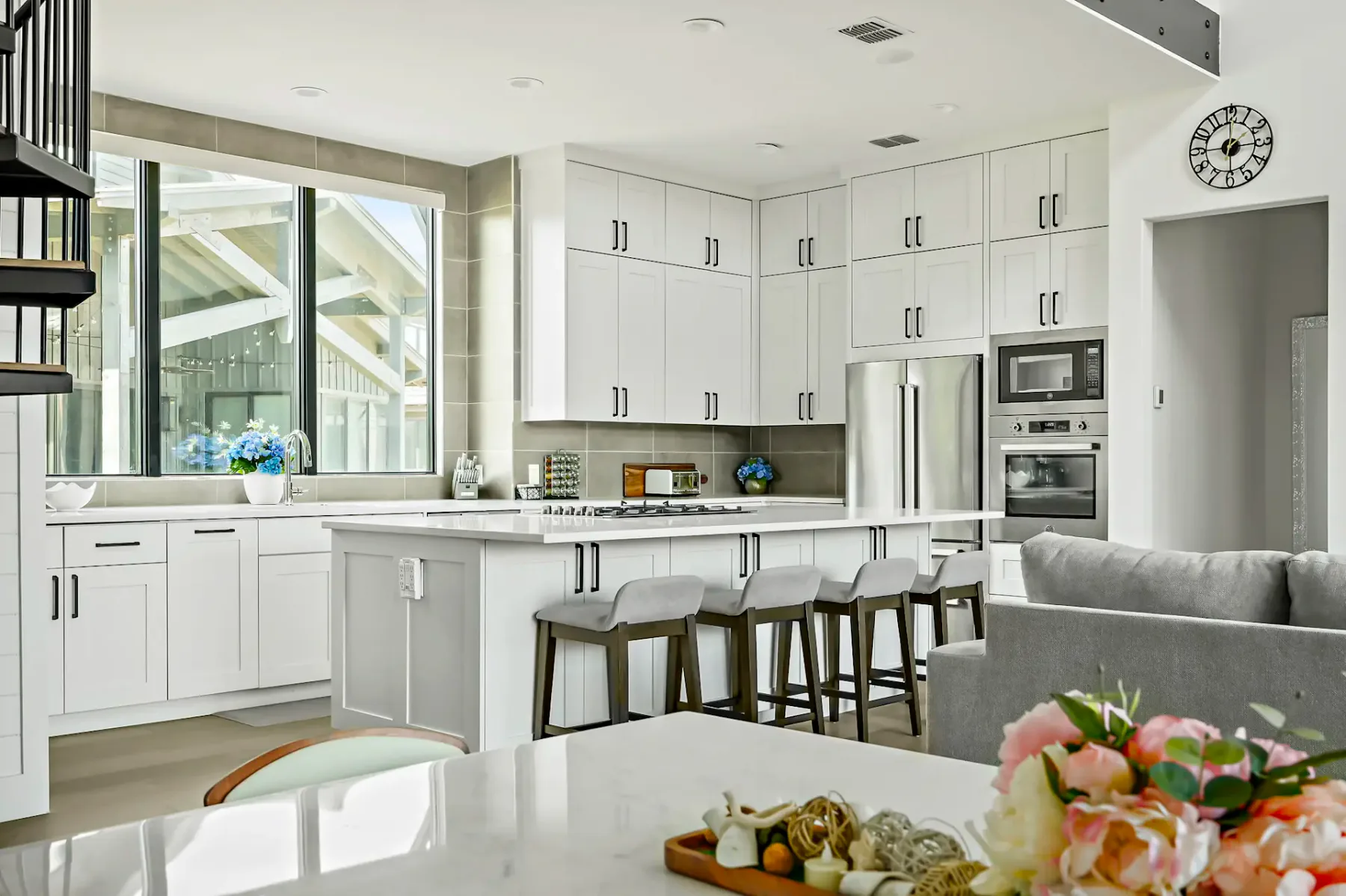 Modern kitchen with white cabinets, center island with bar stools, stainless steel appliances, and large window. Floral decor on the counter.
