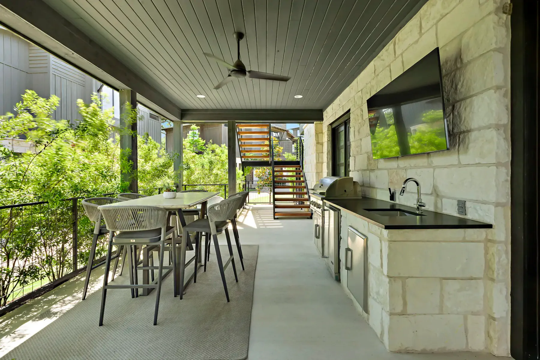 A covered patio with a dining table, chairs, TV, grill, and ceiling fan, overlooks lush greenery, with a staircase in the background.