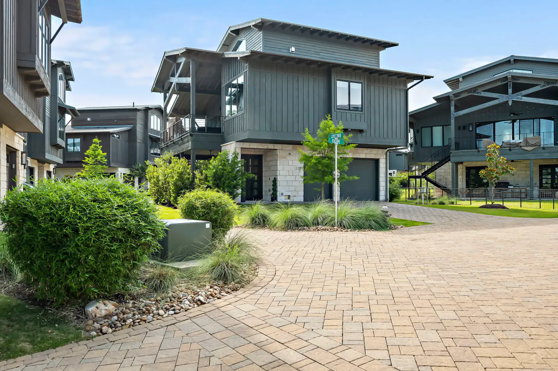 Modern grey houses with large windows, surrounded by greenery and neatly paved driveways, under a clear blue sky.
