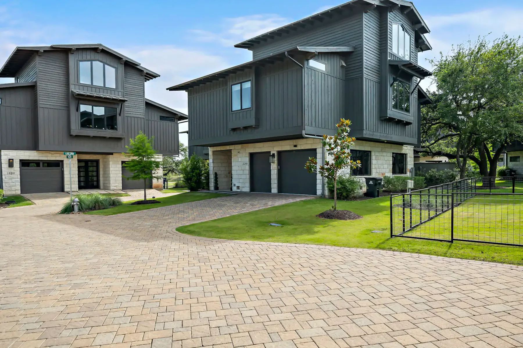 Modern two-story houses with dark siding and stone accents, paved driveways, green lawns, and a black metal fence under a blue sky.
