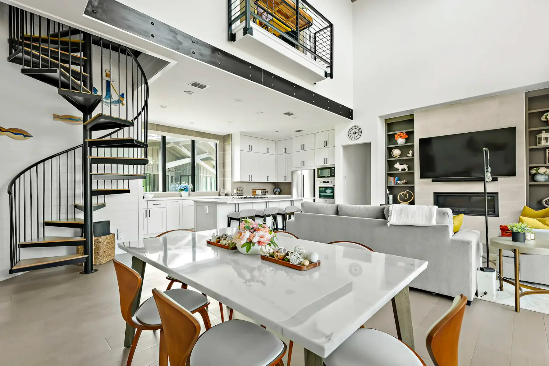 Modern open-concept living area with spiral staircase, white kitchen, glass dining table, gray sofas, and wall-mounted TV near a fireplace.