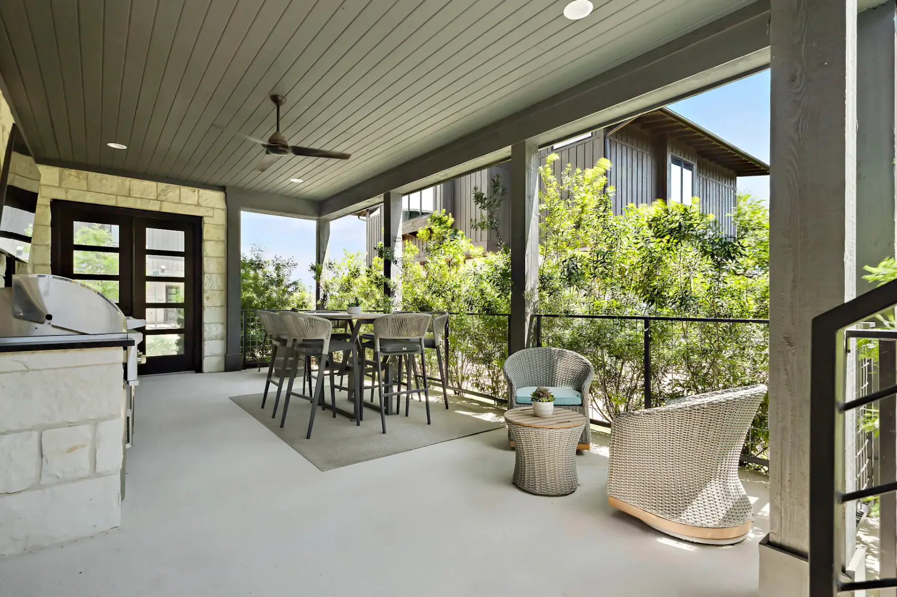 Modern covered patio with dining table, chairs, and wicker seating. Ceiling fan overhead and lush greenery outside. Bright and inviting space.