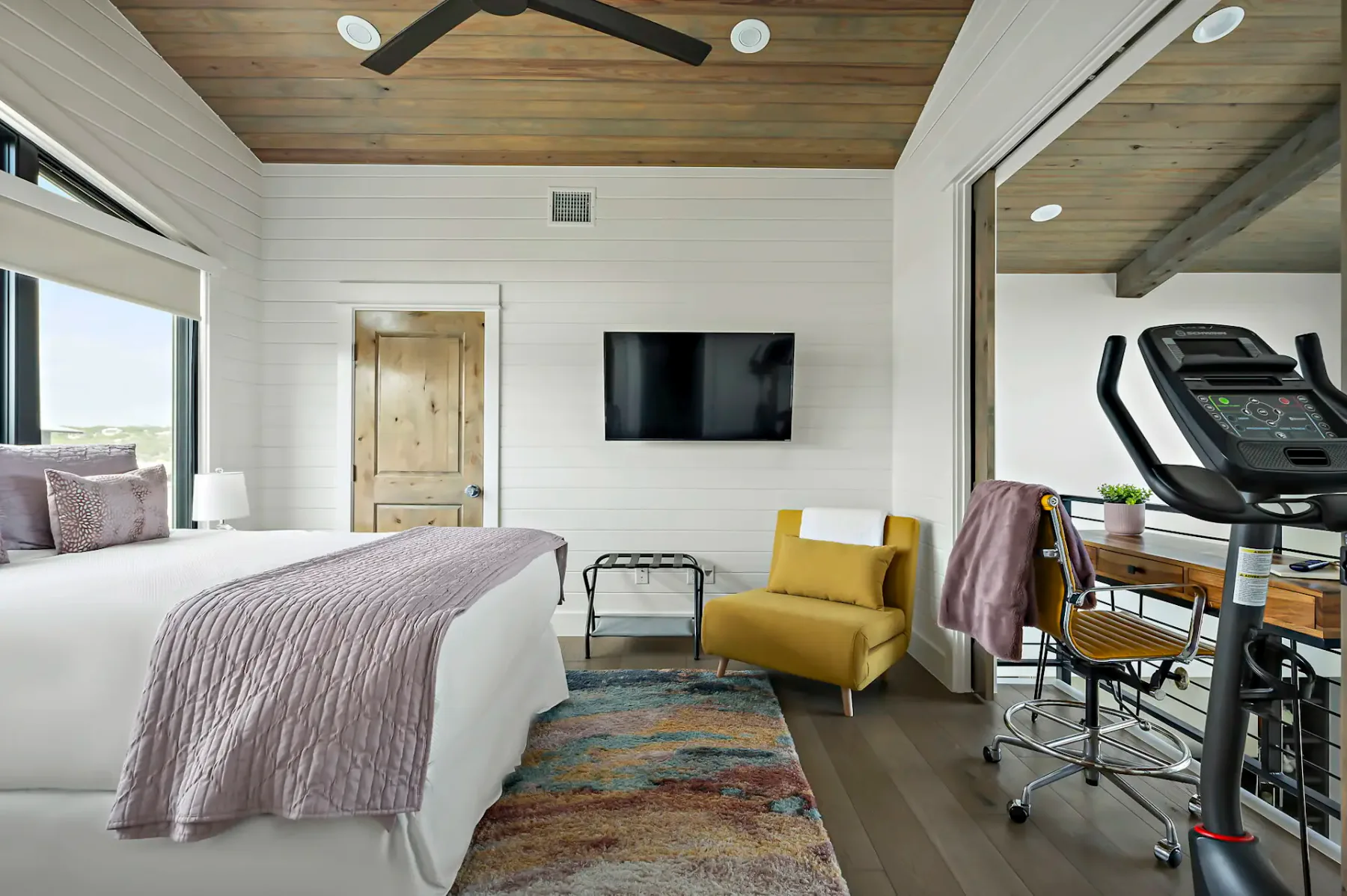 A bedroom with a bed, television, yellow chair, and exercise equipment.