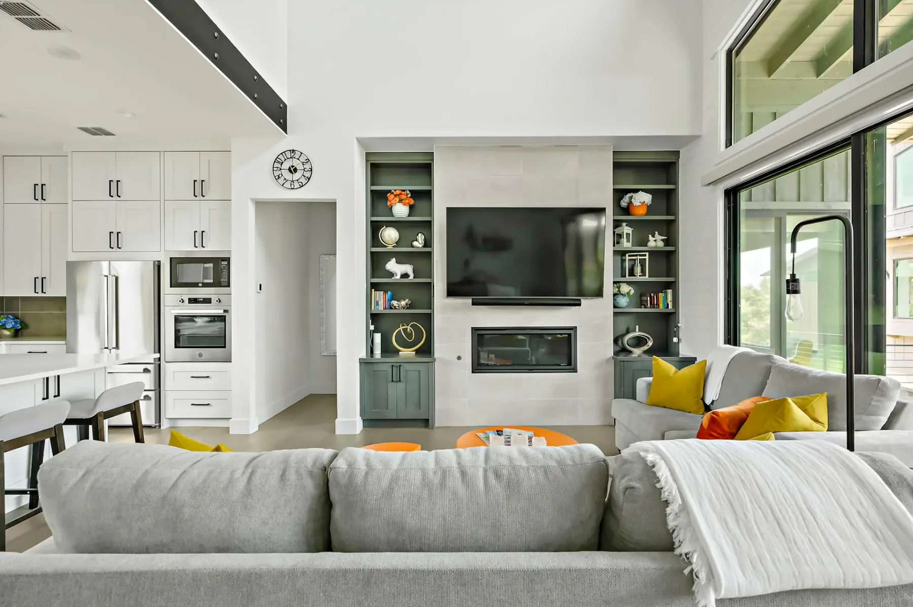 Modern living room with a wall-mounted TV, a fireplace, shelves, light gray sofa, and large windows. Neutral tones with pops of orange and yellow.