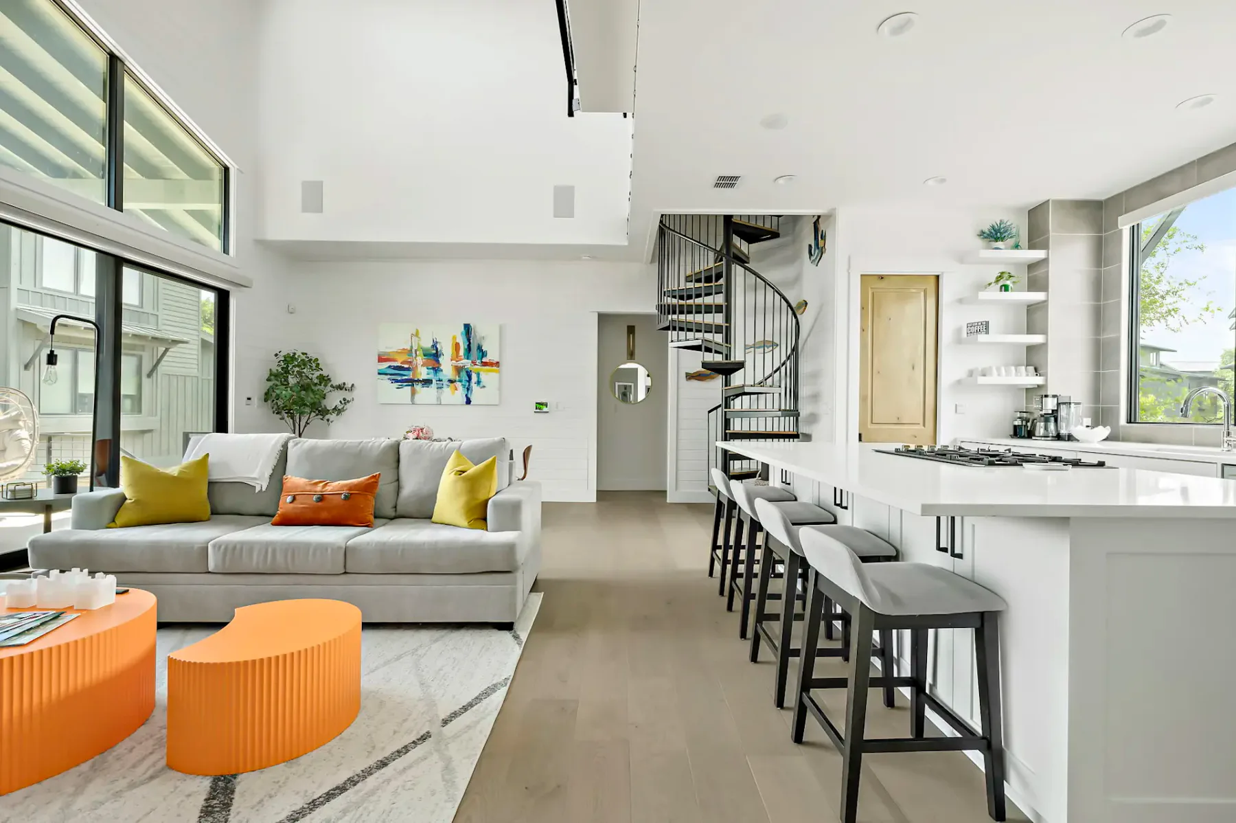 Modern living room with gray sofa, colorful cushions, orange coffee tables, spiral staircase, and kitchen island with stools. Bright and airy space.