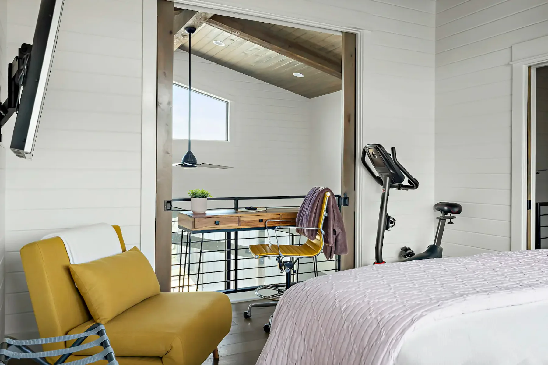 A bedroom with a bed, yellow seating, an exercise bike, and a desk in the adjoining loft area.