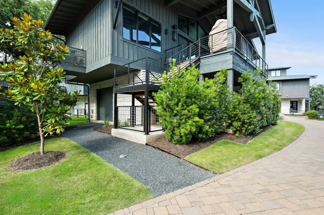 Modern two-story house with gray siding and elevated porch, surrounded by green shrubs, on a neatly paved and landscaped yard.