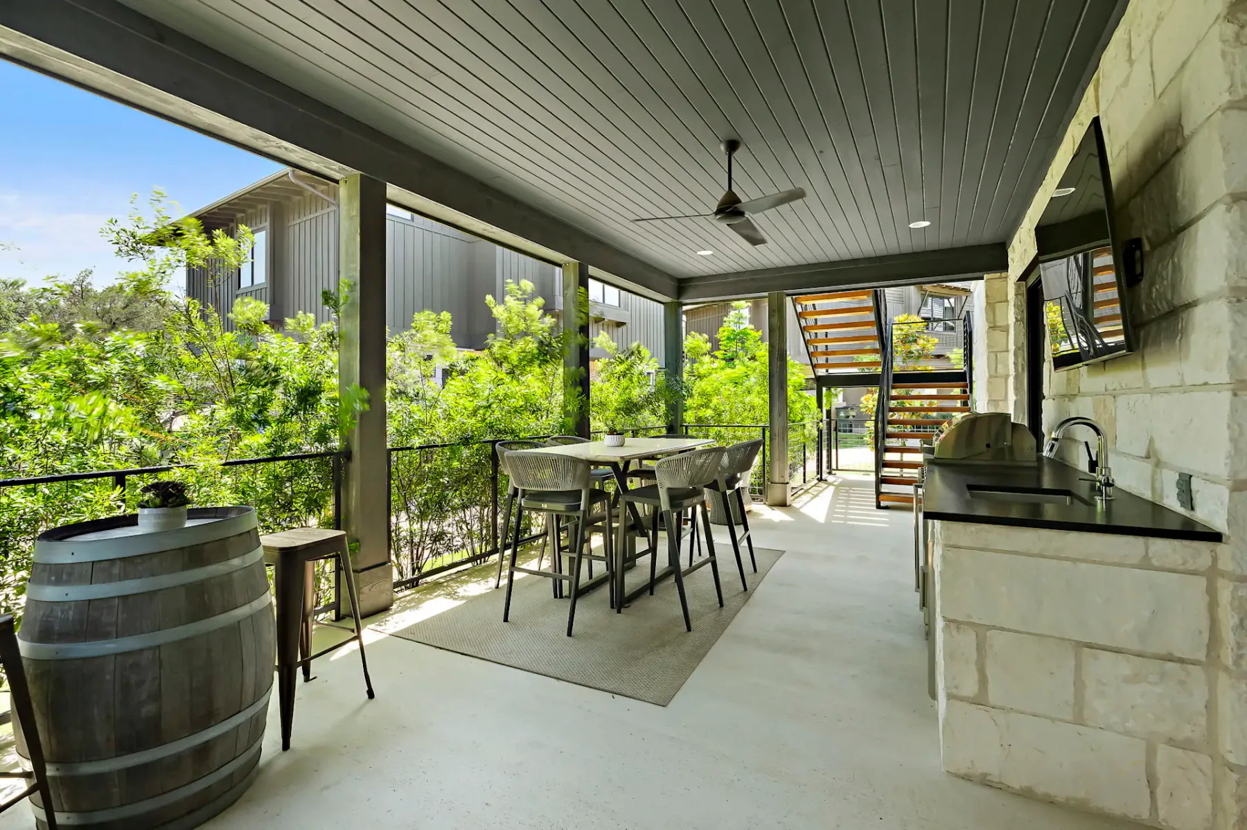 Covered outdoor patio with a ceiling fan, high-top table and chairs, wine barrel, and built-in grill, surrounded by greenery.
