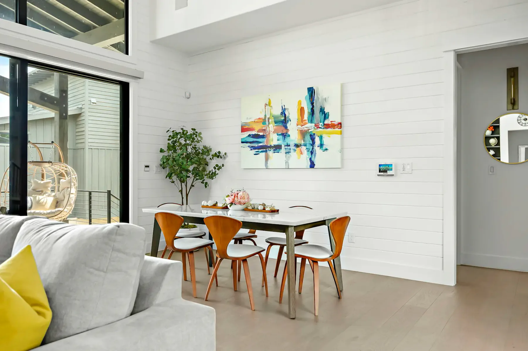 Modern dining area with a white table, wooden chairs, and colorful abstract art on the wall. Large windows provide natural light.