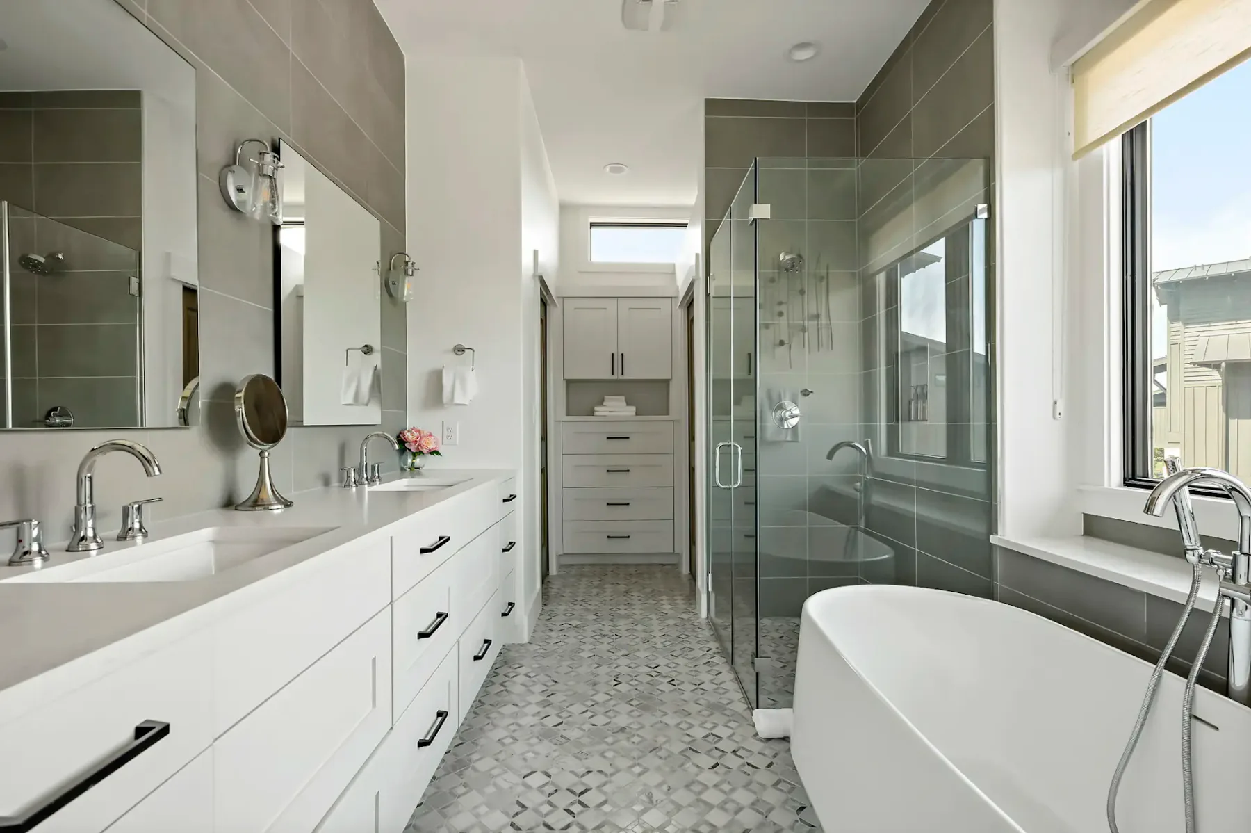 Modern bathroom with a white vanity, double sinks, glass-enclosed shower, freestanding tub, and large window. Gray tile accents the walls and floor.