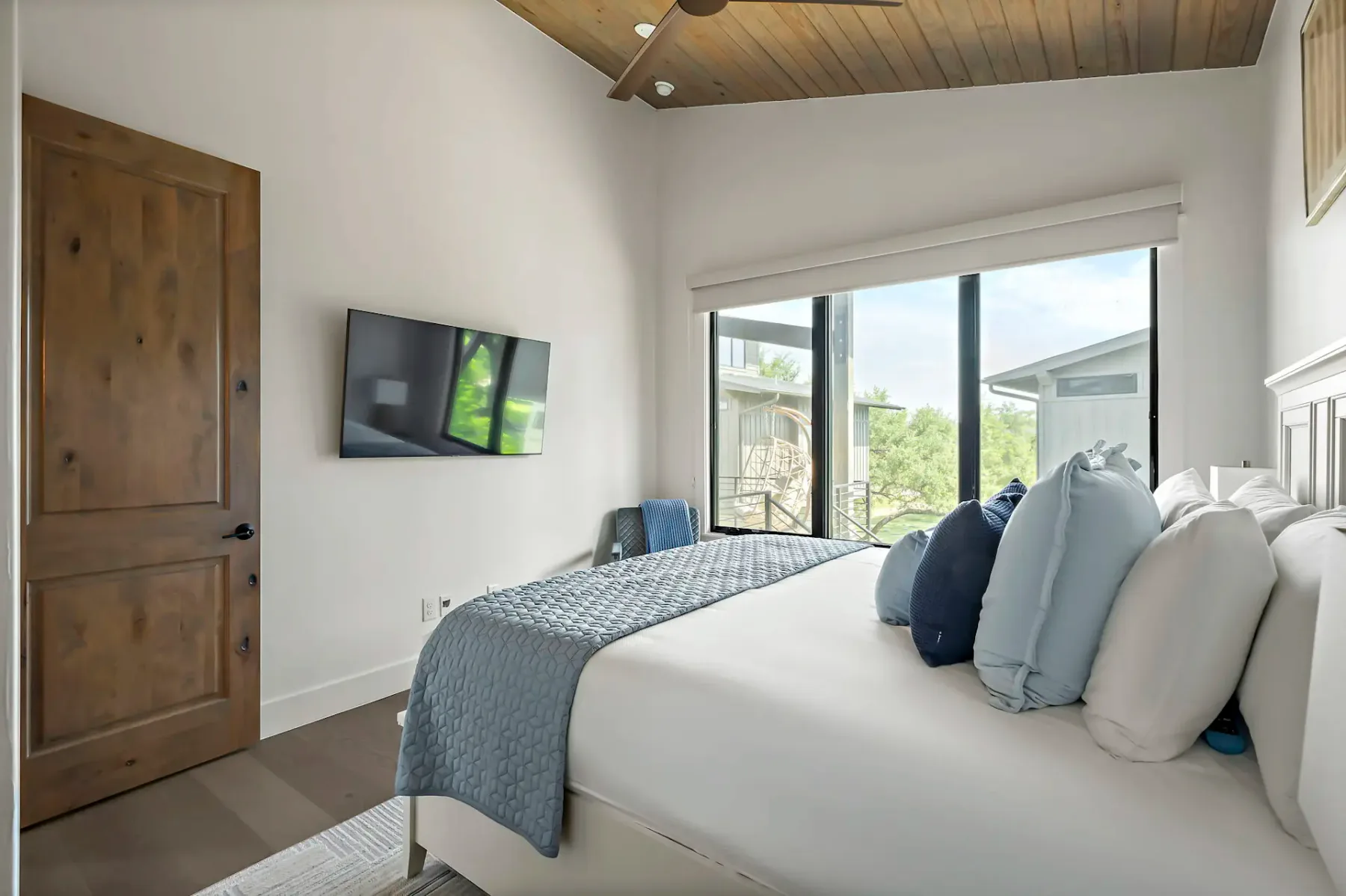 Cozy bedroom with a large bed, blue and white pillows, a wall-mounted TV, wooden door, and a large window offering a view outside.