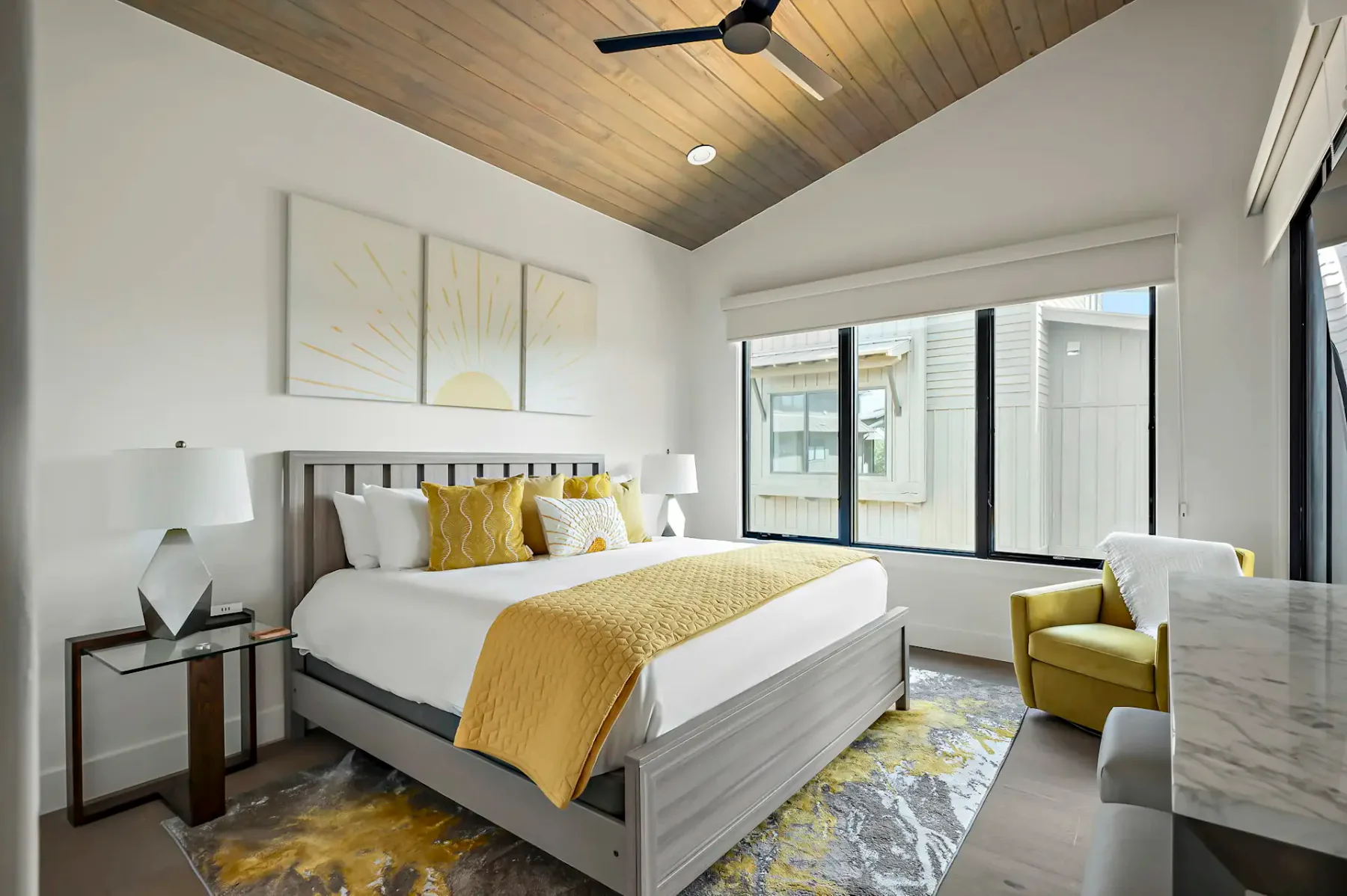 Modern bedroom with a wooden ceiling, large bed with yellow accents, abstract art, yellow armchair, and a window with an outdoor view.