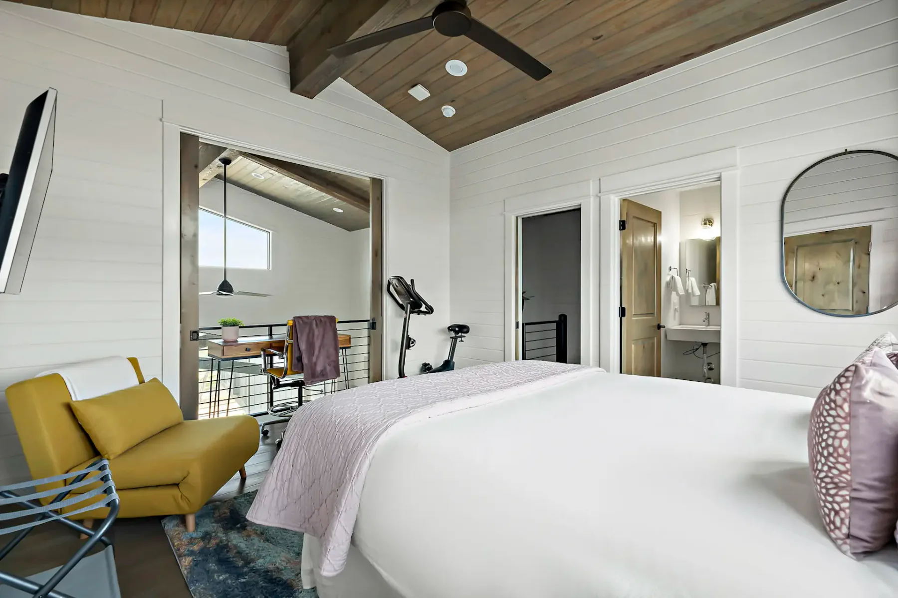 A bedroom with a large bed, yellow seating, and an exercise bike near the doorway.