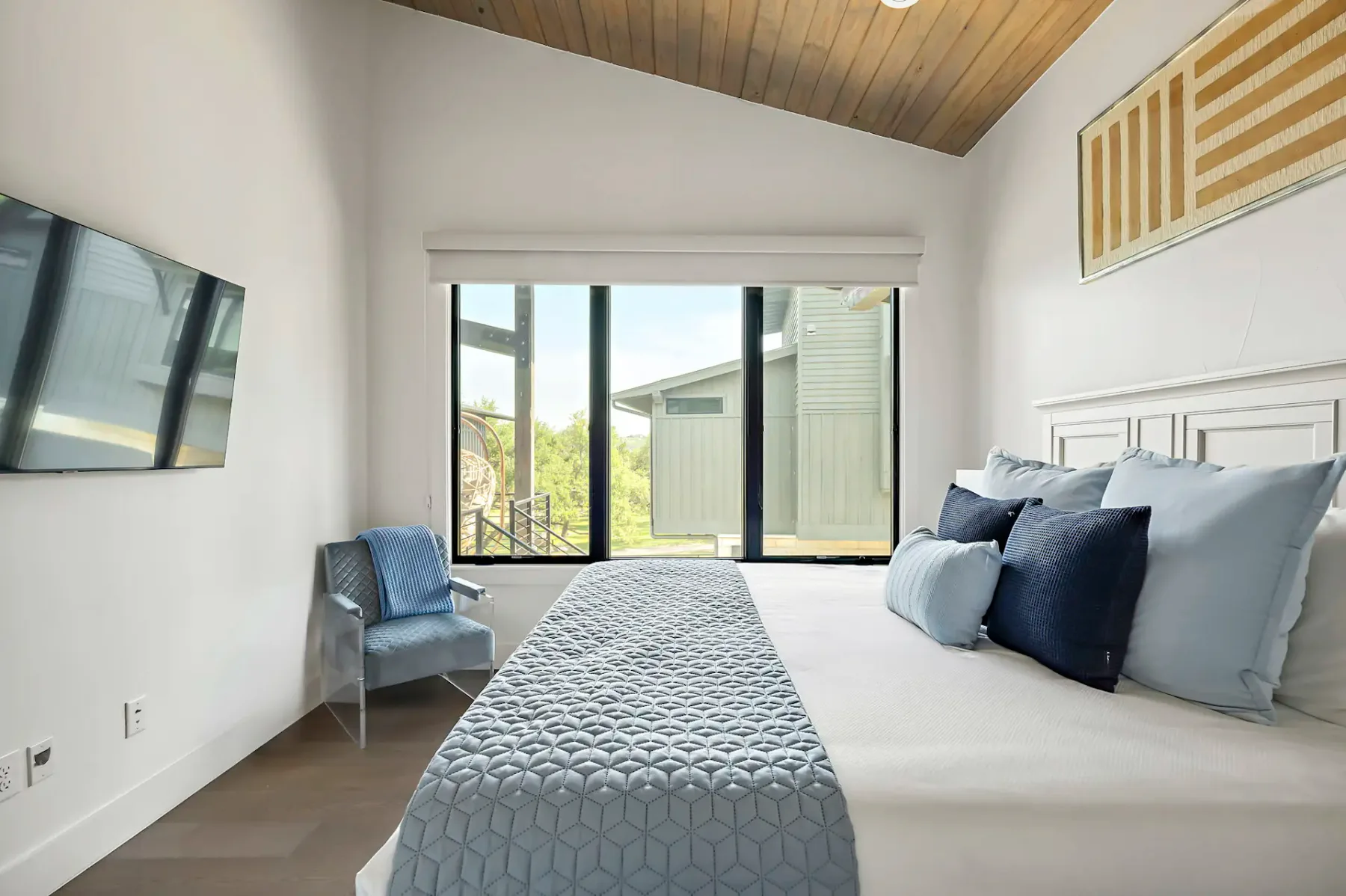 Modern bedroom with large windows, a bed with blue and white bedding, a blue chair, and a wall-mounted TV. Ceiling has wood paneling.
