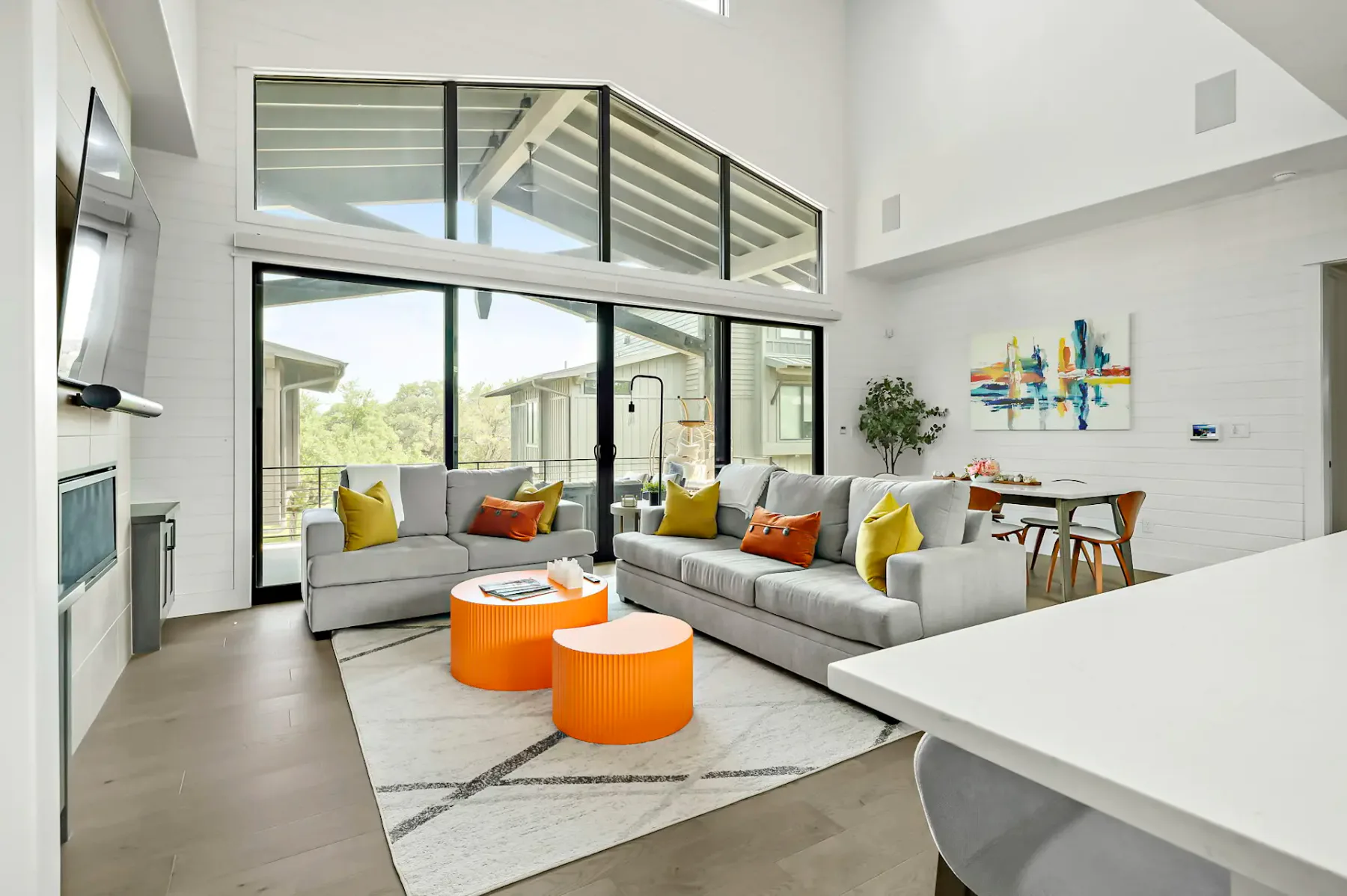 Modern living room with gray sofas, colorful cushions, orange coffee tables, large windows, abstract wall art, and a dining area in the background.