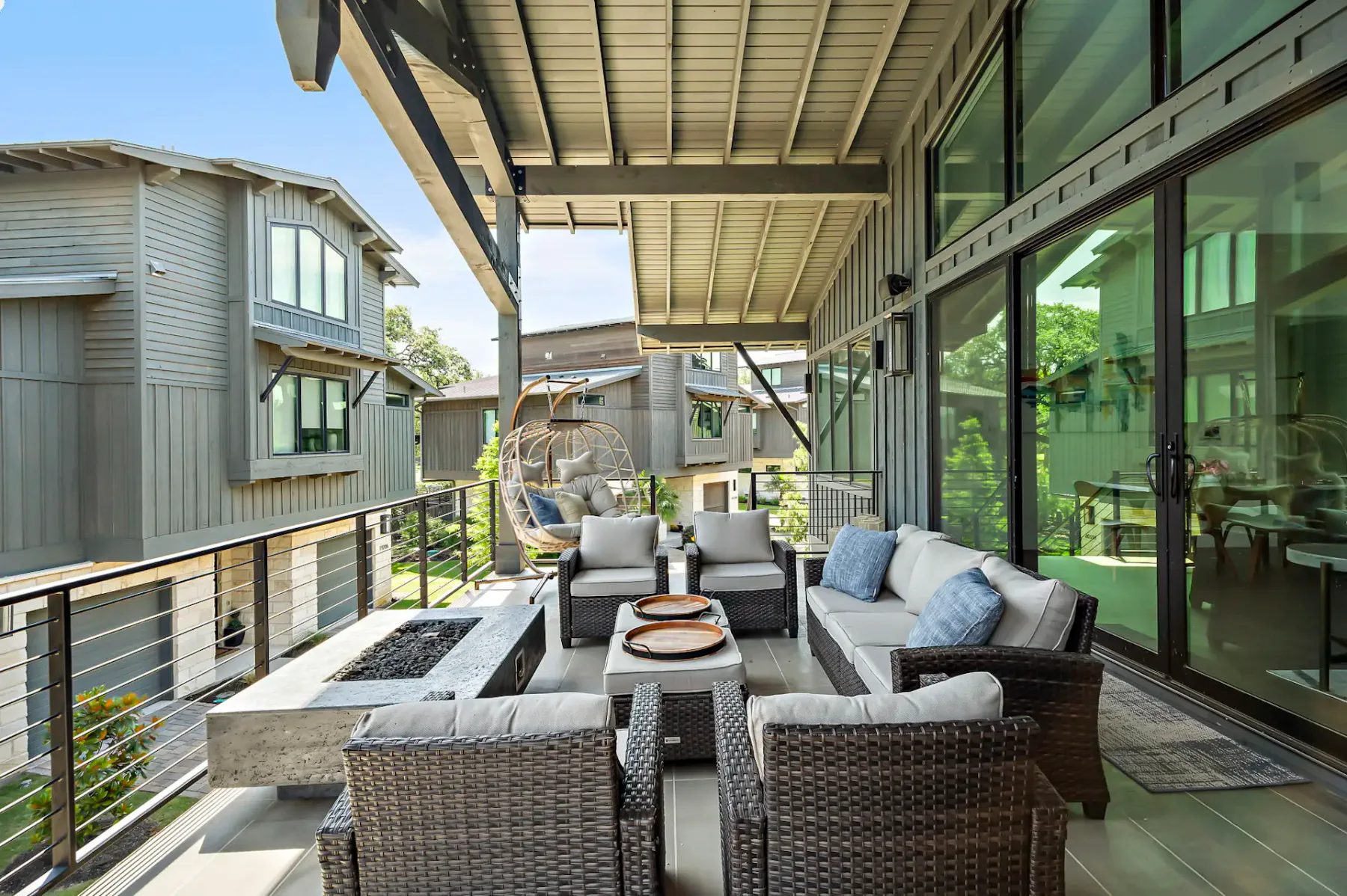 Modern patio with wicker furniture, cushions, a fire pit table, and a hanging chair, overlooking adjacent contemporary houses.