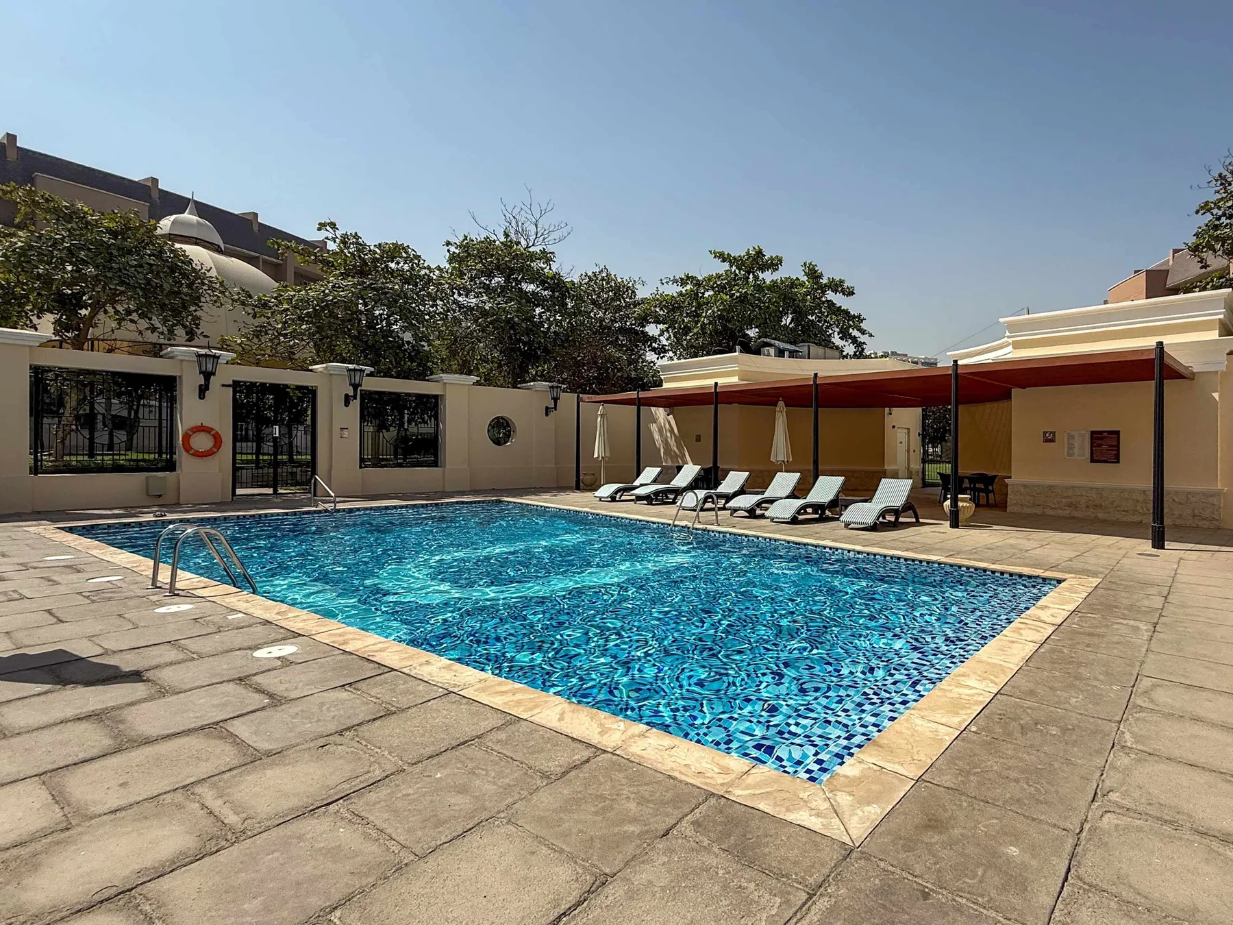 Una piscina al aire libre está junto a una terraza pavimentada y asientos a la sombra.