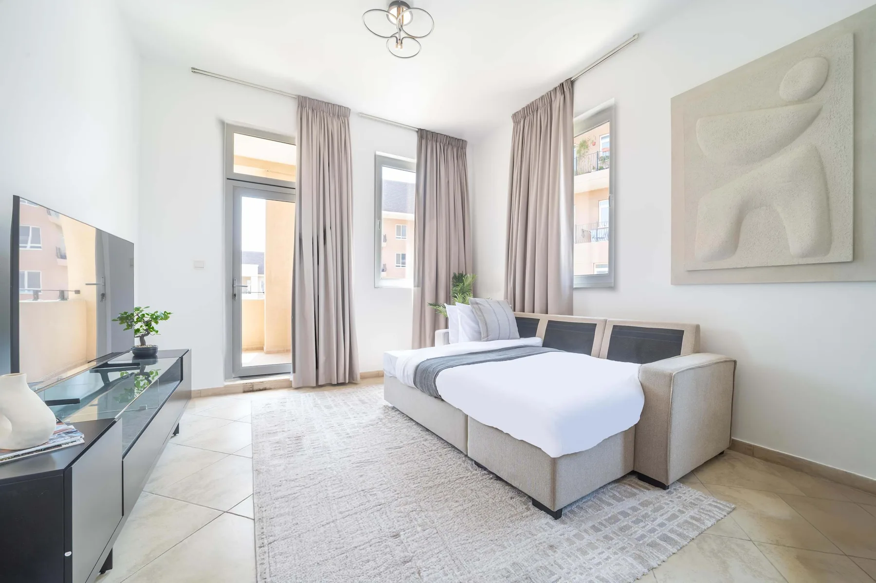 Un dormitorio luminoso con una cama, ventanas altas y cortinas claras.