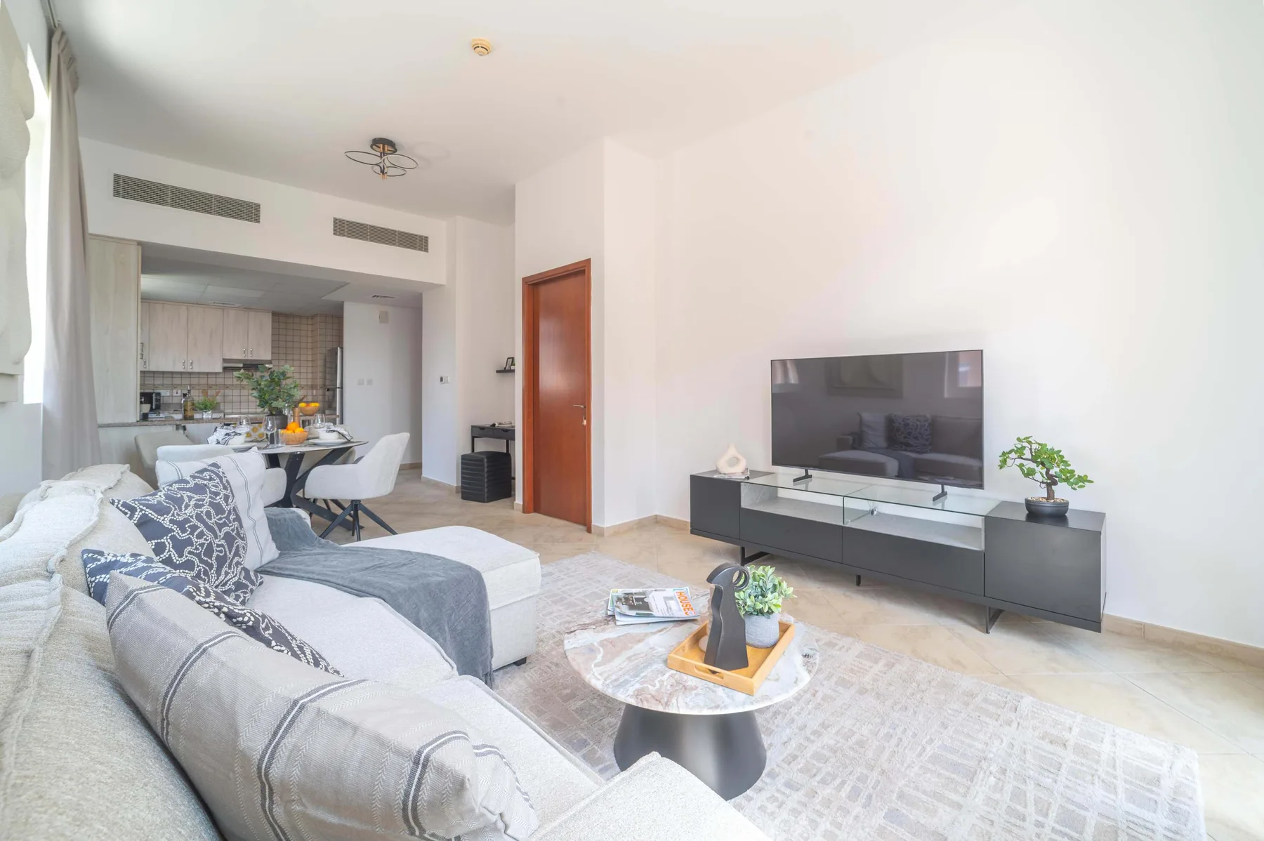 A bright living room with seating, a television, and an open kitchen in the background.