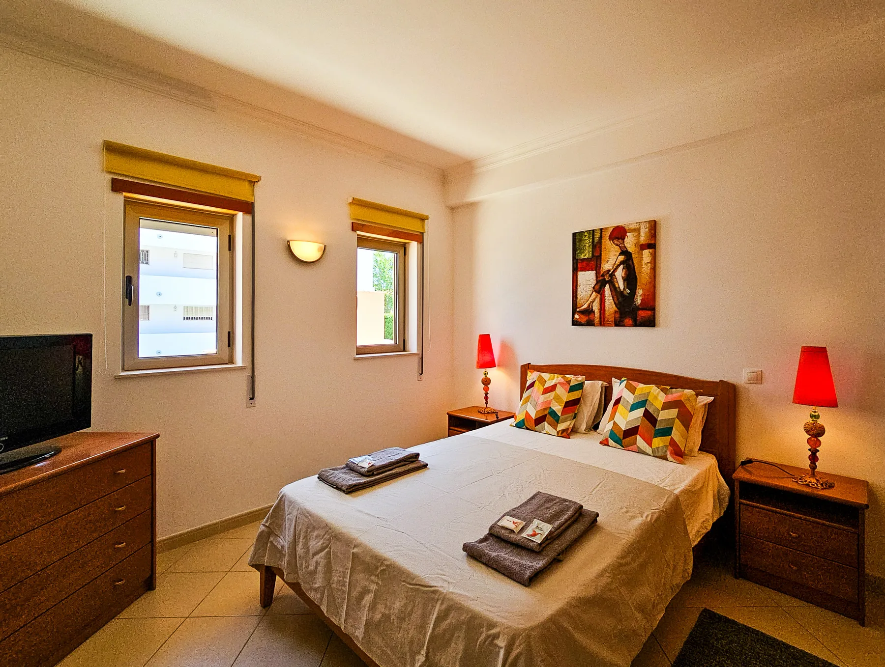 A bedroom with a double bed, colorful pillows, and two windows.