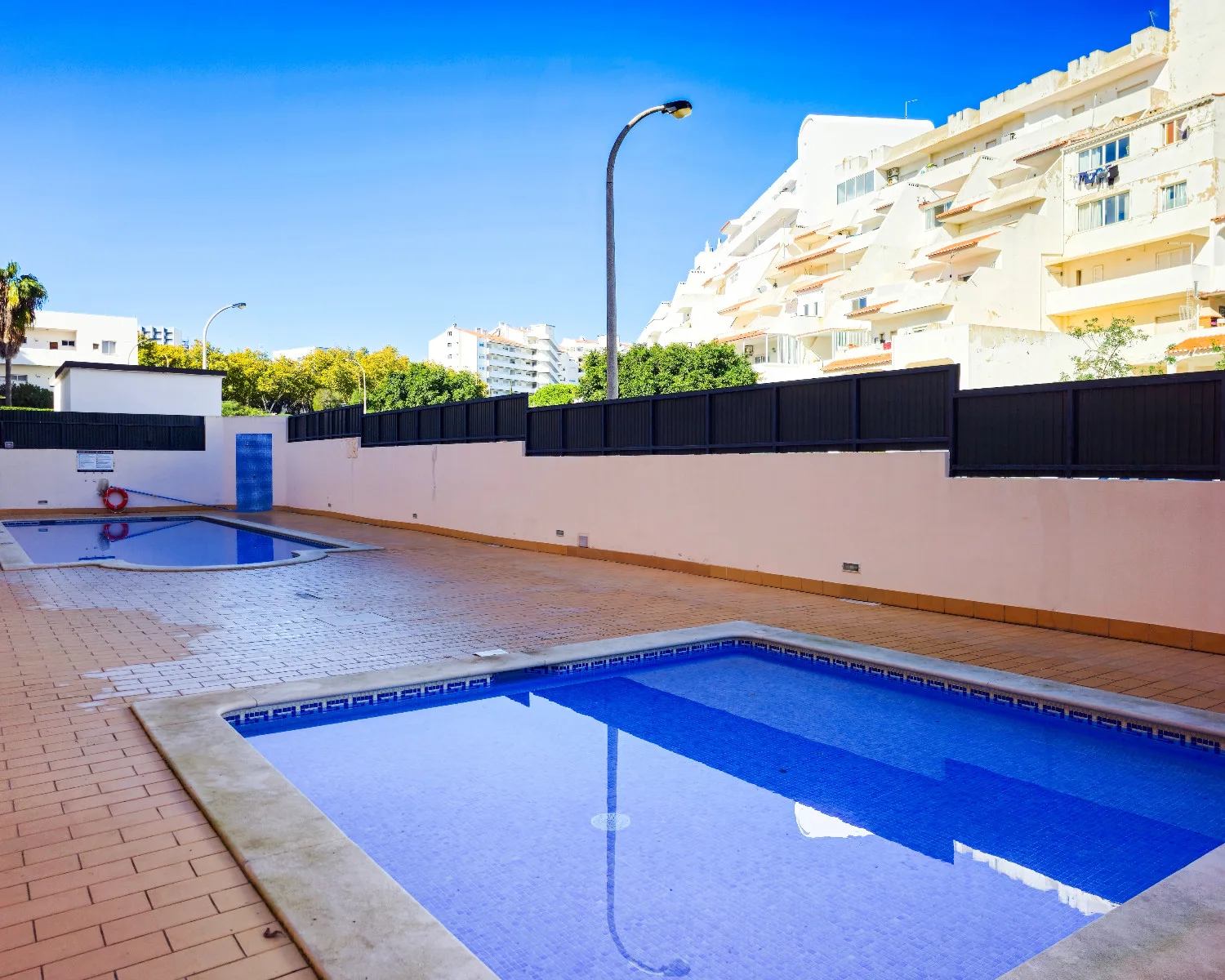 An outdoor swimming pool area sits beside sunlit apartment buildings.