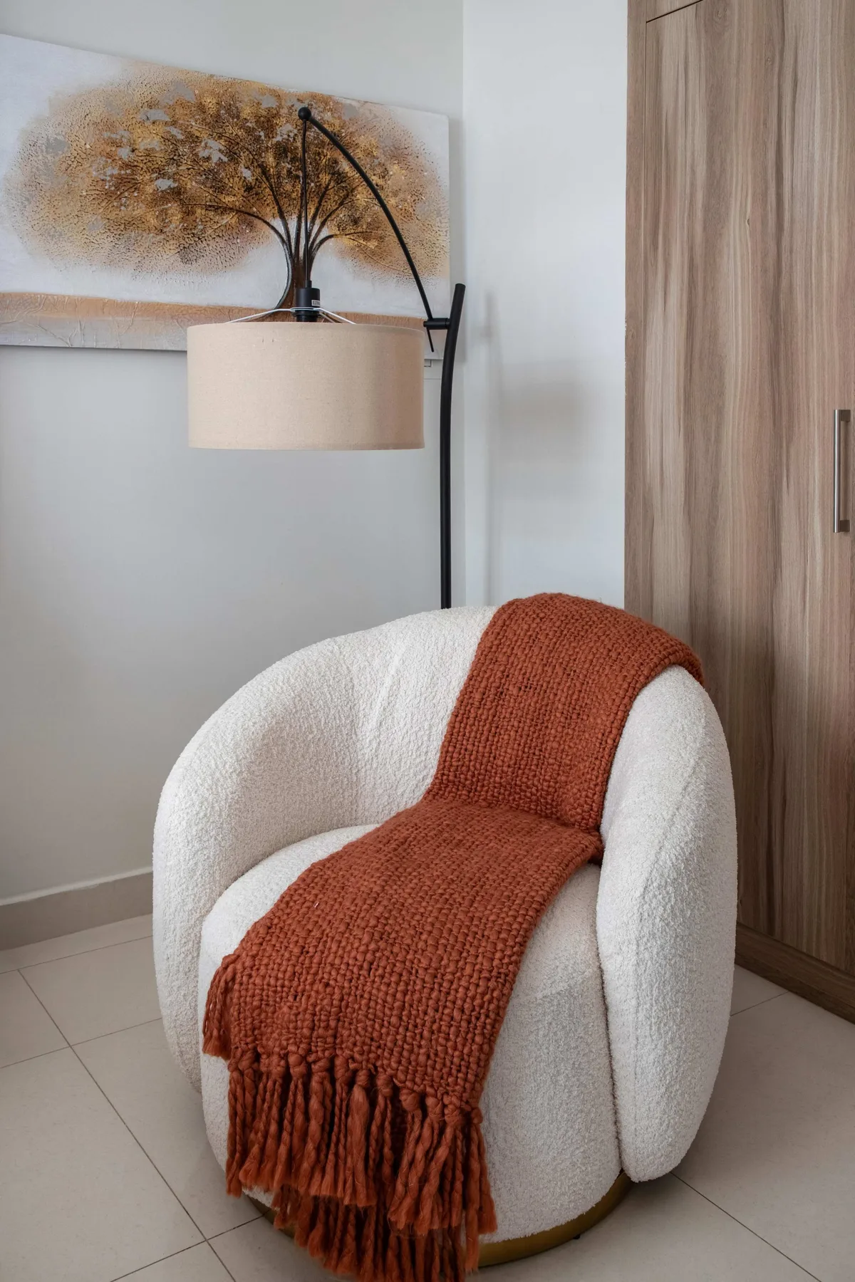 Un sillón blanco con una manta color óxido está junto a una lámpara de pie.
