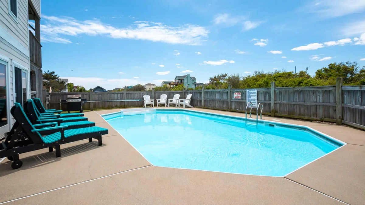 An outdoor swimming pool sits beside a house, with lounge seating along the deck.