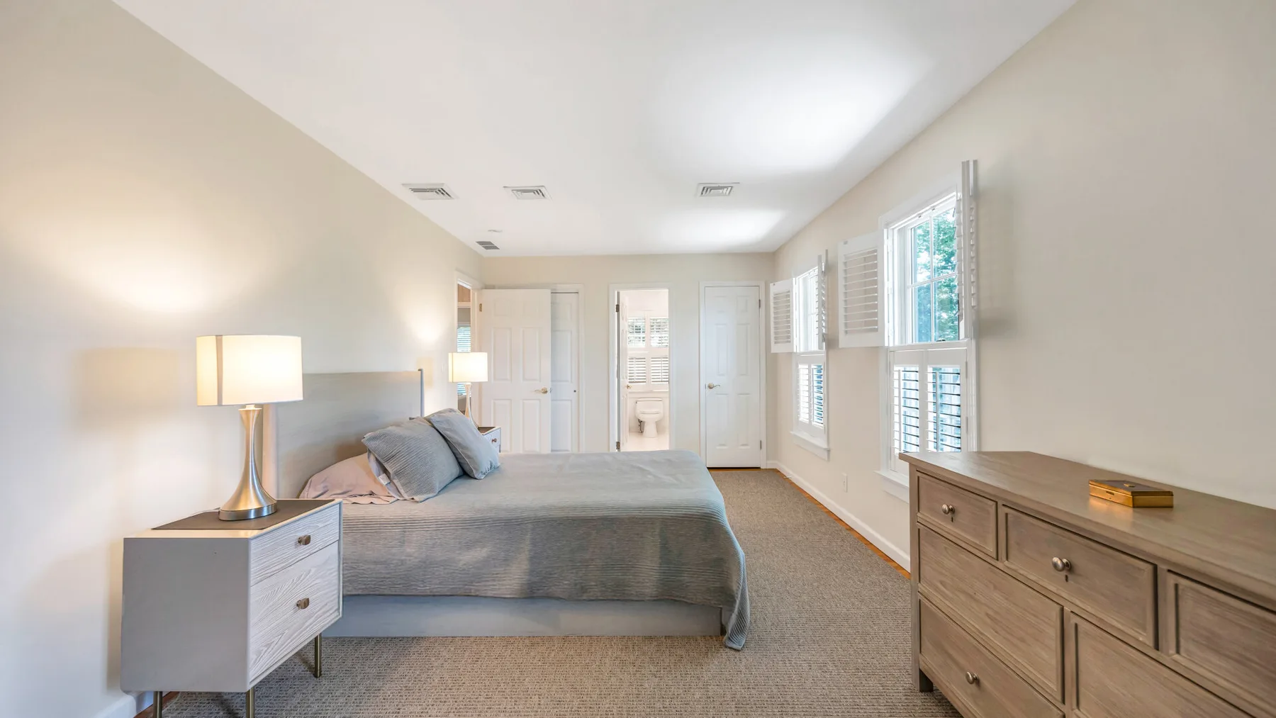 Un dormitorio luminoso con una cama, una lámpara de noche y ventanas amplias.