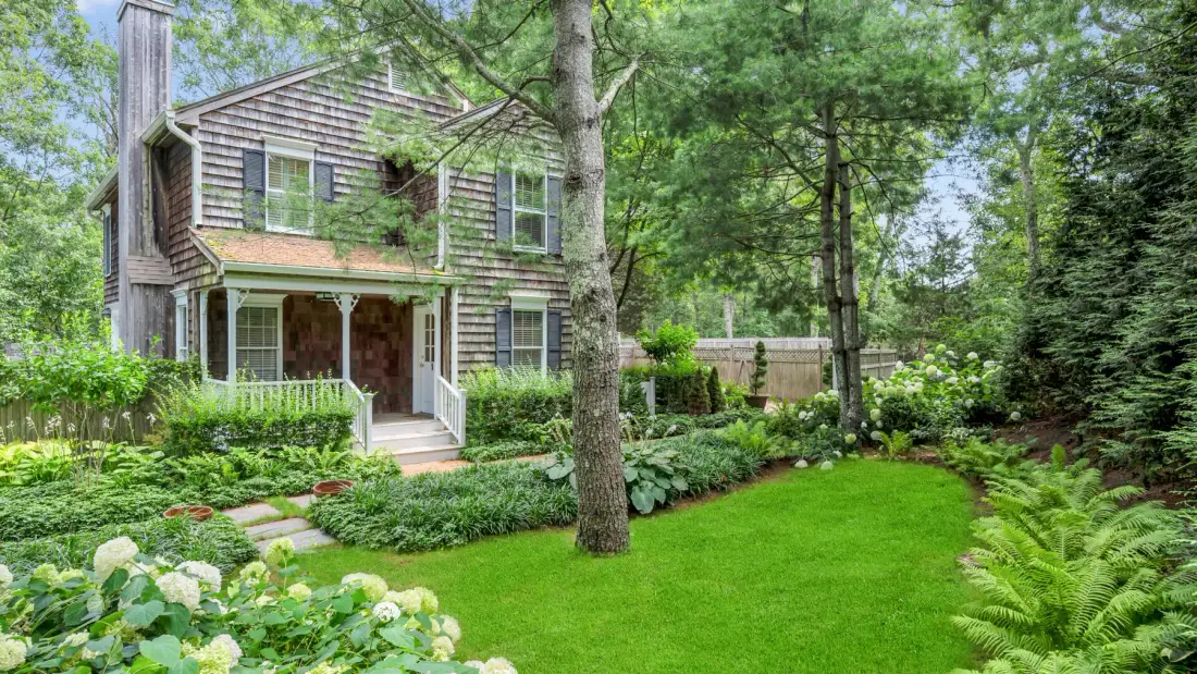 A two-story house stands beside a green lawn and leafy garden.