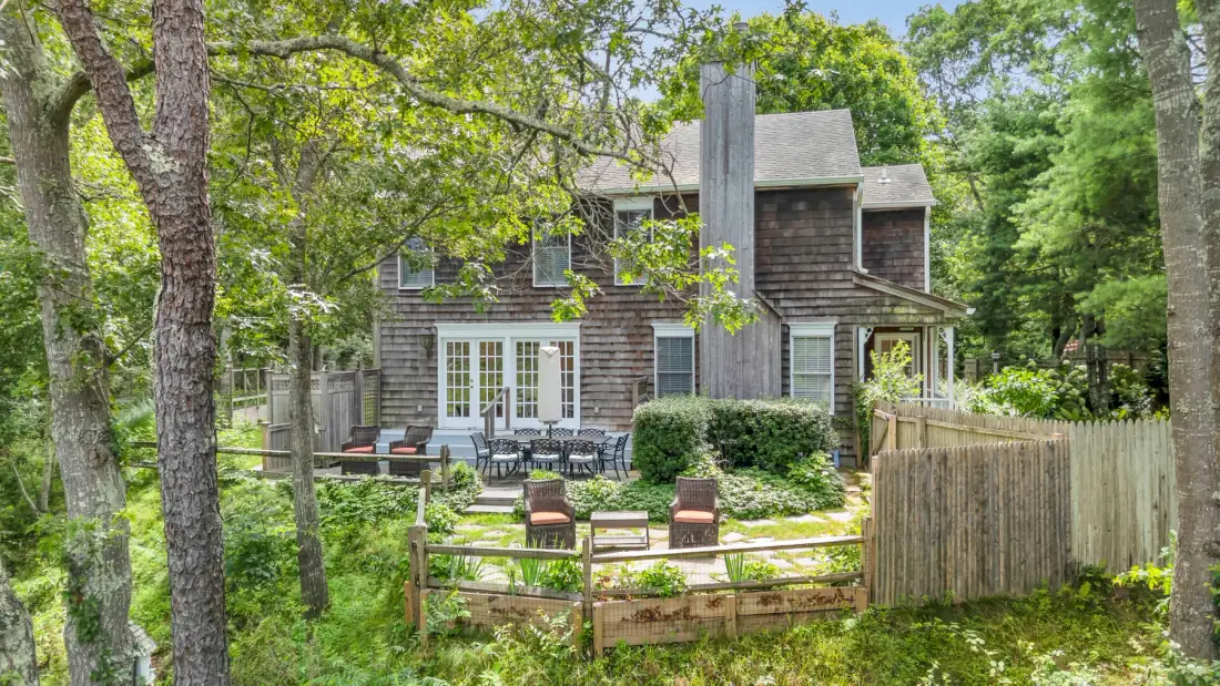 Una casa rodeada de árboles, con jardín cercado y asientos al aire libre.