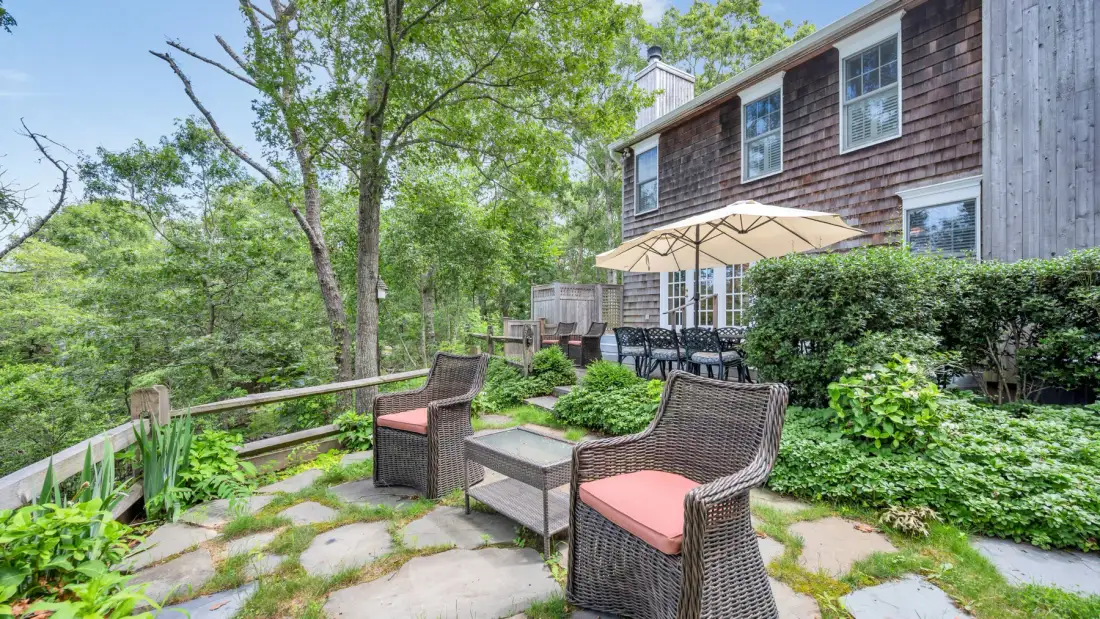 An outdoor patio with cushioned seating beside a house and leafy garden.