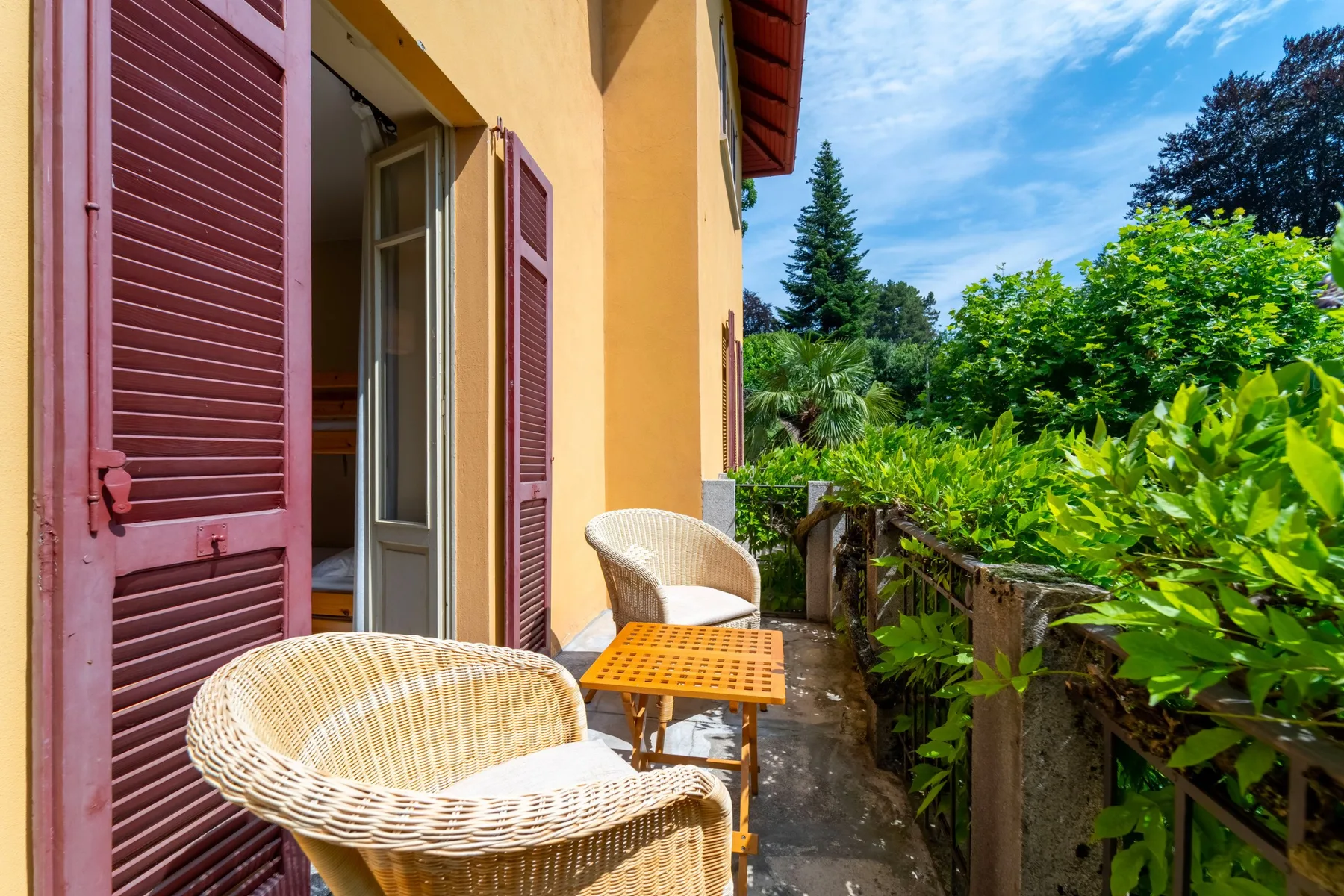 A sunny balcony with wicker seating, an open doorway, and leafy greenery.