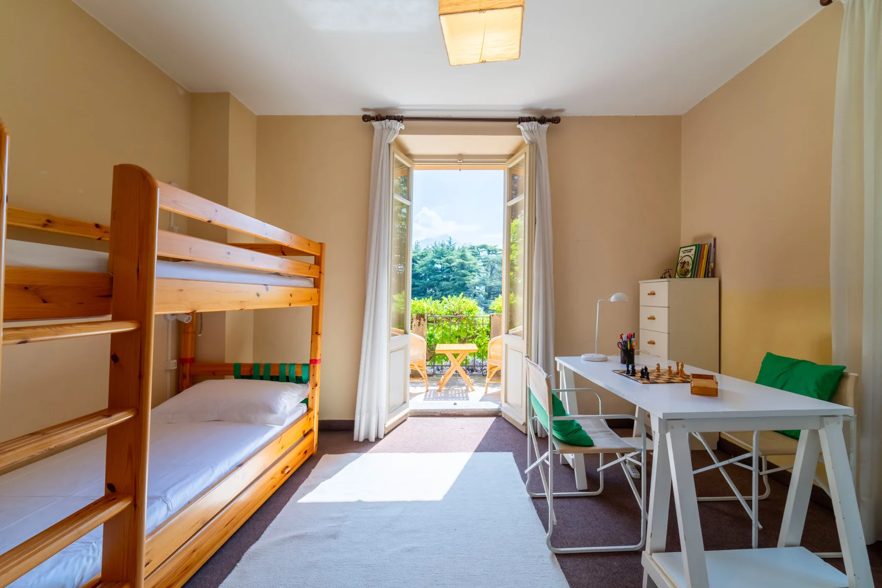 A bright bedroom with bunk beds, a table, and an open window overlooking greenery.