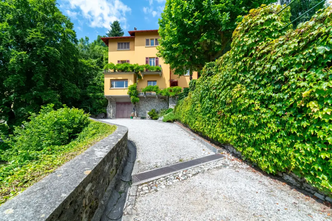 A gravel driveway curves toward a yellow house amid lush greenery.