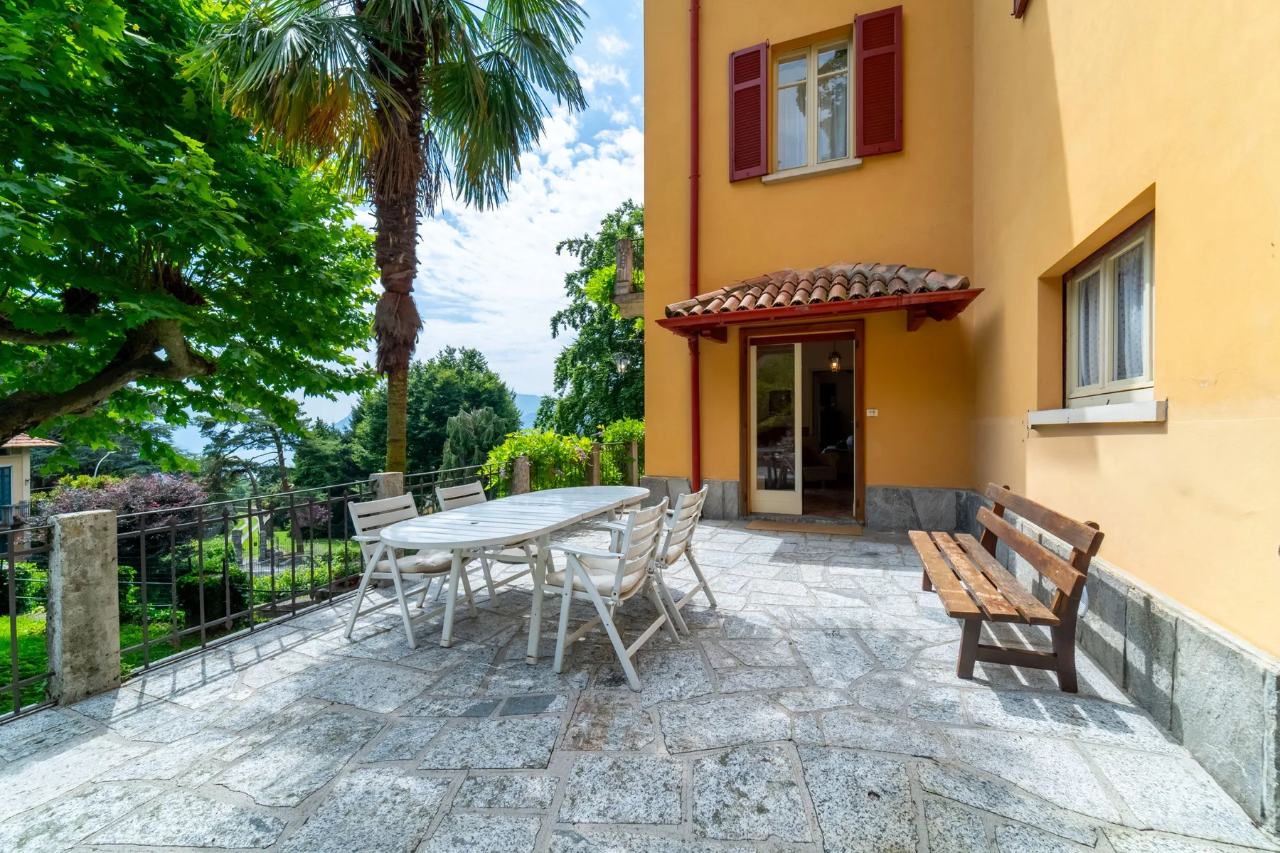 A stone terrace beside a yellow building has outdoor seating and leafy trees.